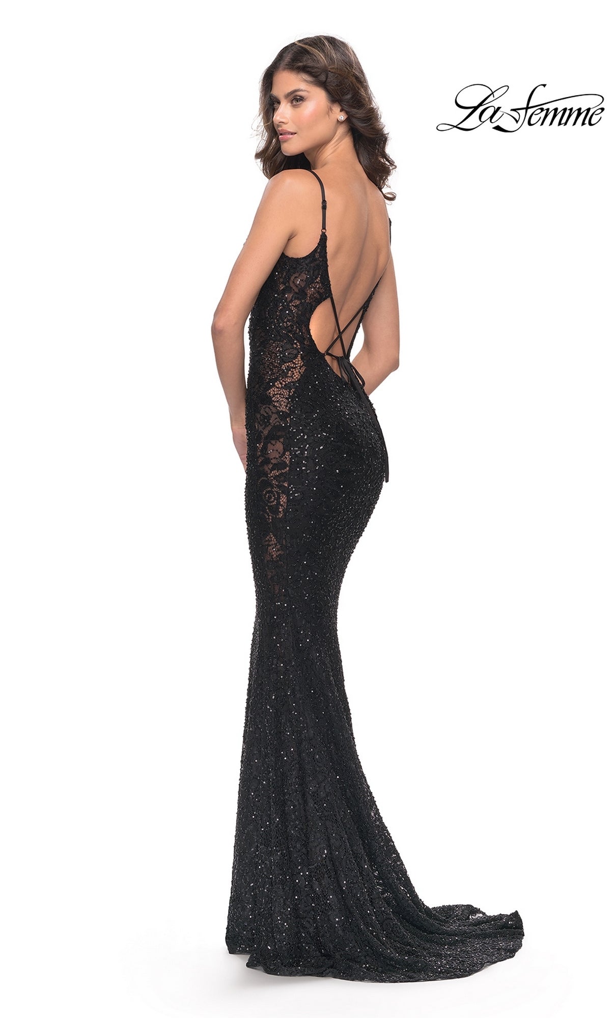 A woman wears the La Femme Beaded Long Black Lace Prom Dress 31257, a sleeveless, backless, floor-length gown with thin straps and intricate lace details, standing to the side against a white background.